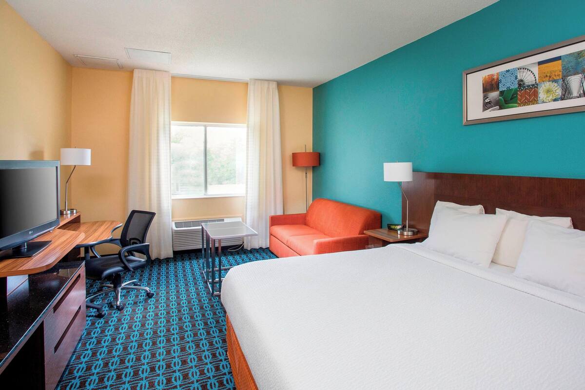 A hotel room with a bed, a desk with a computer, a chair, a sofa, and a TV.