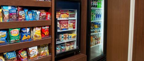 A snack shelf stocked with various chips and snacks, a mini-fridge with beverages, and a small refrigerator with hot food items.