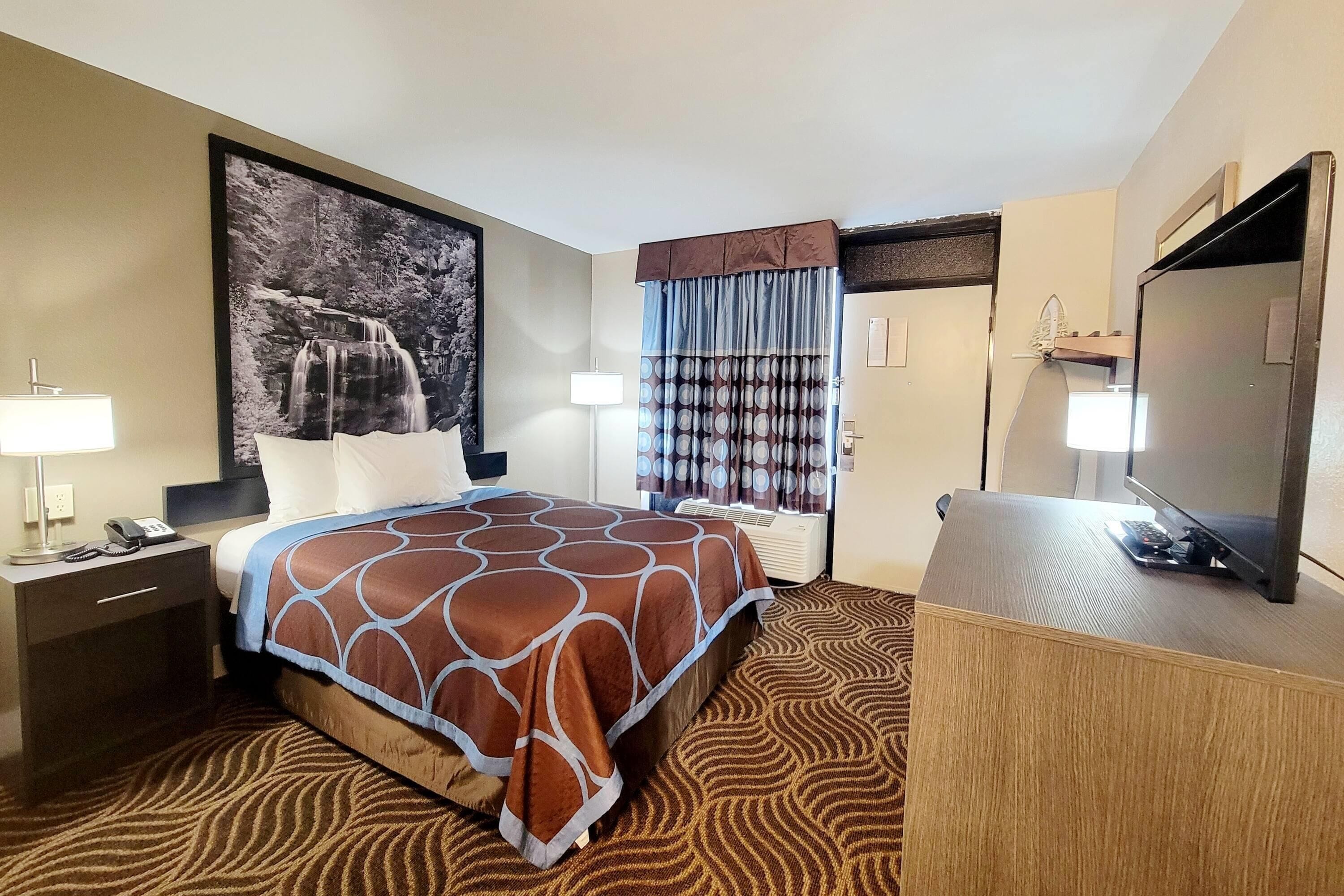 A hotel room with a bed, a desk with a lamp, a television, and a large wall-mounted picture.