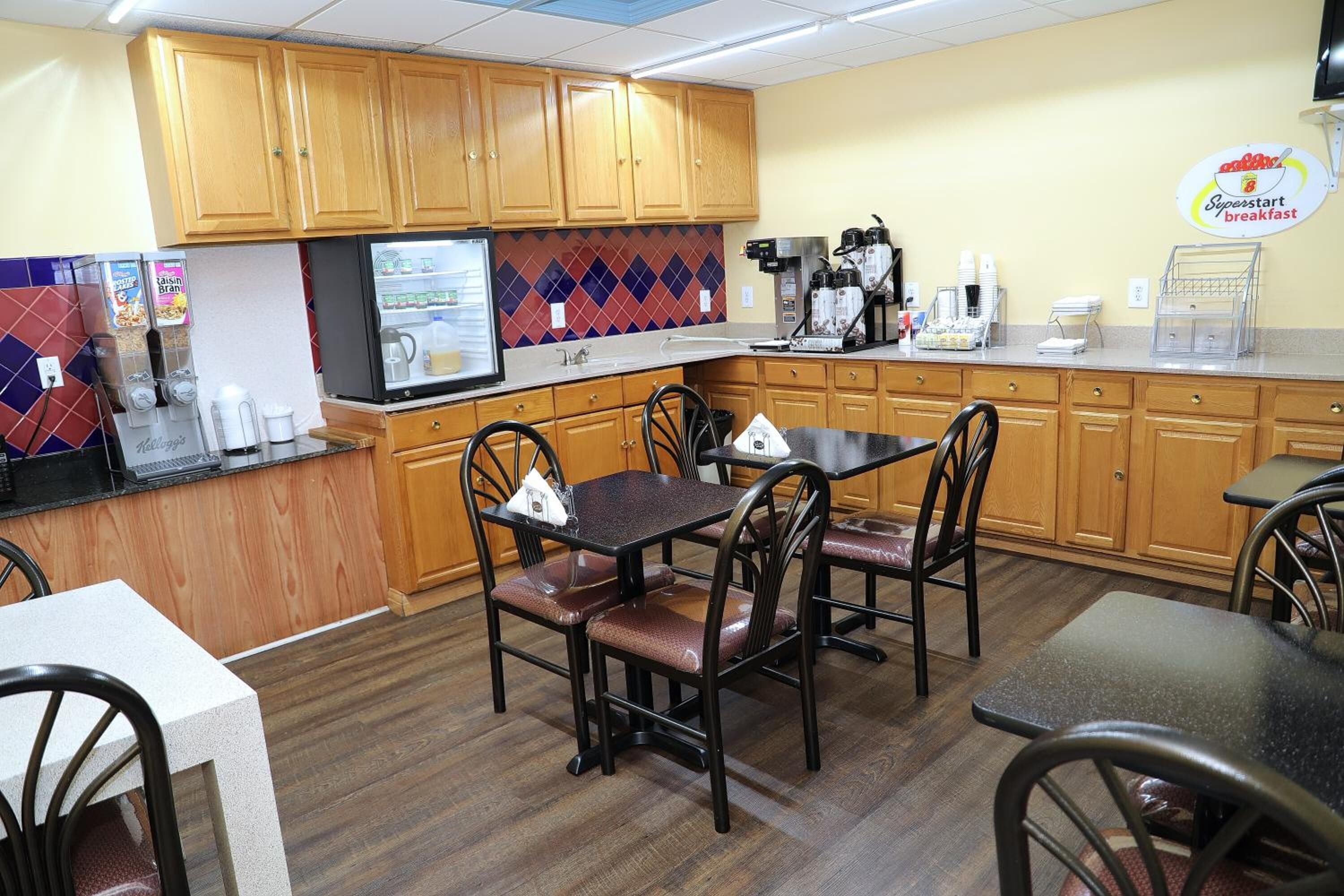 A small, well-lit kitchen with wooden cabinets, a refrigerator, a coffee station, and a dining area with chairs and tables.