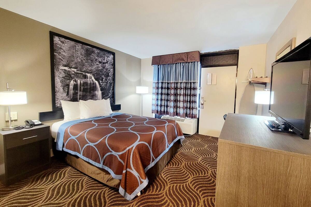 Una habitación de hotel con cama, un escritorio con computadora, teléfono, lámpara y un amplio ventanal con cortinas.