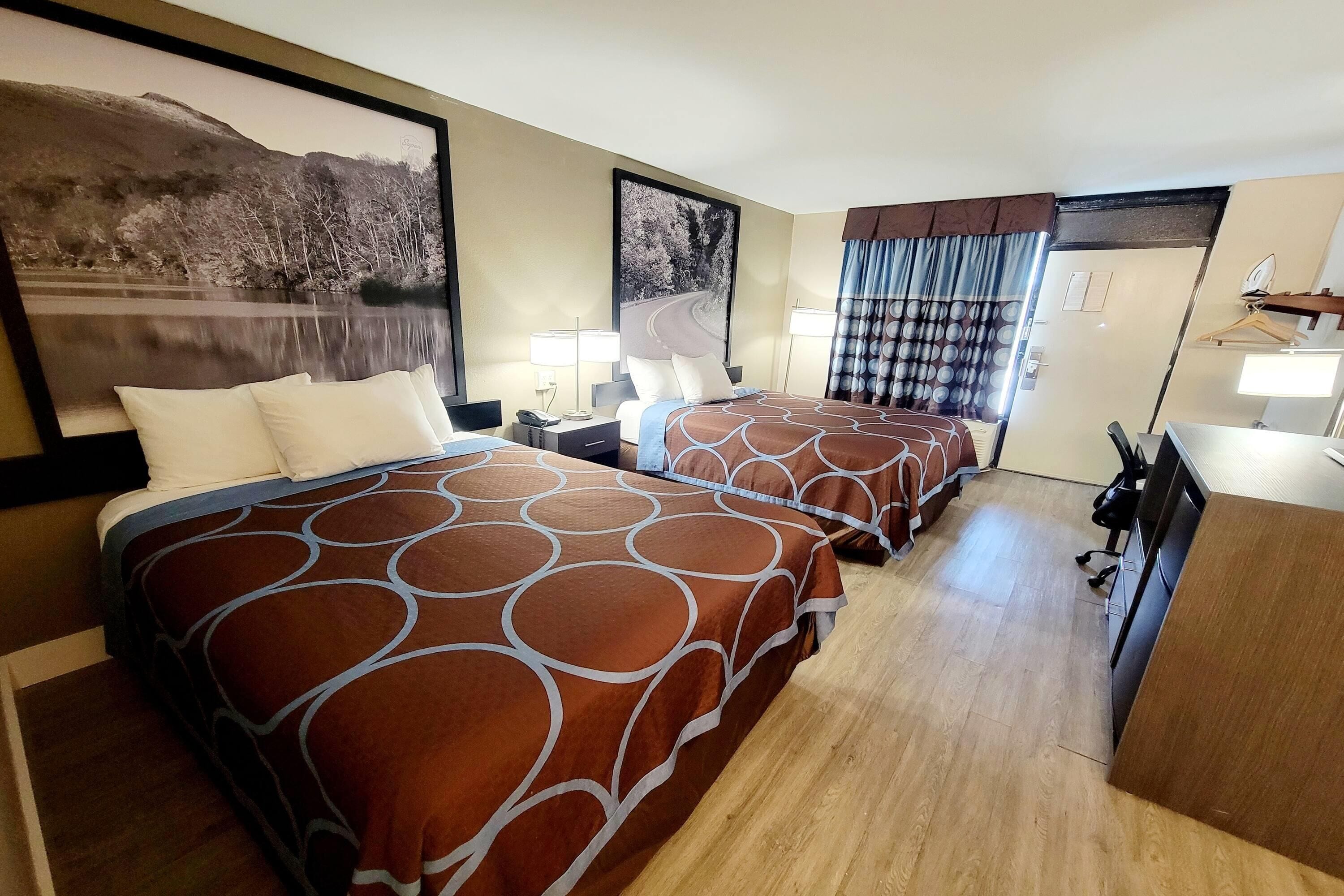 A hotel room with two beds, a desk, a chair, a lamp, and a large framed picture on the wall.