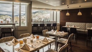 Un restaurante con vista a la ciudad, mesas dispuestas con servilletas y cubiertos, y lámparas colgantes.