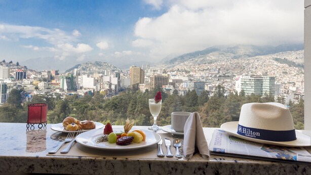 Una mesa de comedor con un plato de comida, una copa de vino y un sombrero, con vistas a un paisaje urbano con montañas al fondo.