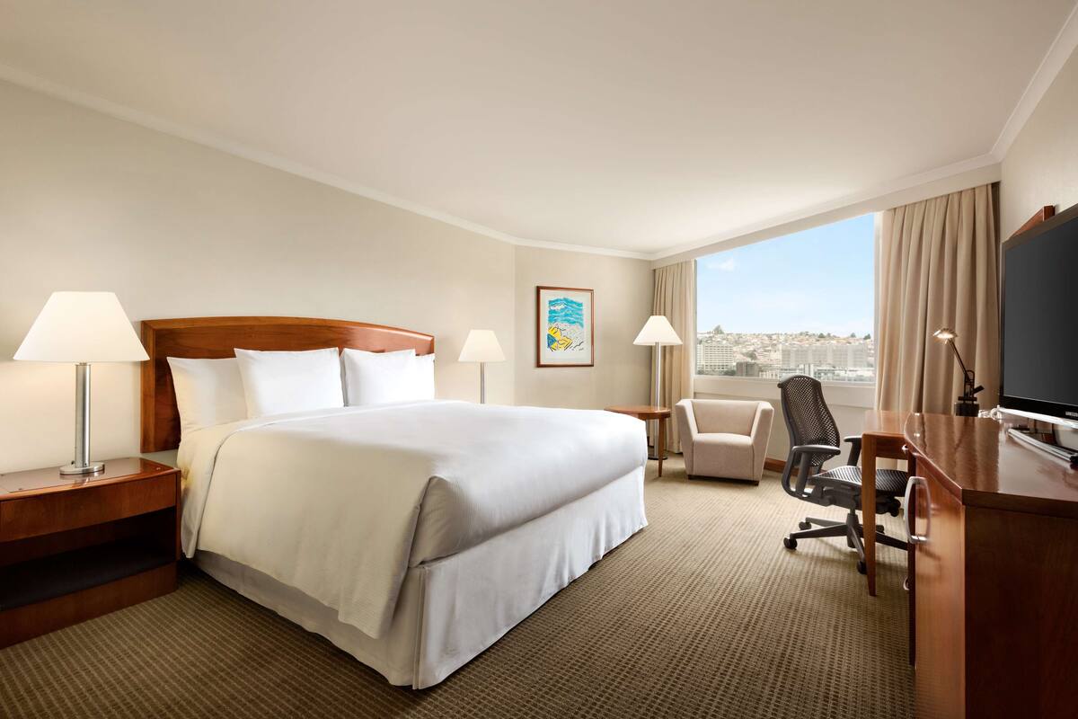 A hotel room with a large bed, two bedside tables with lamps, a desk with a chair, a television, and a window with a city view.