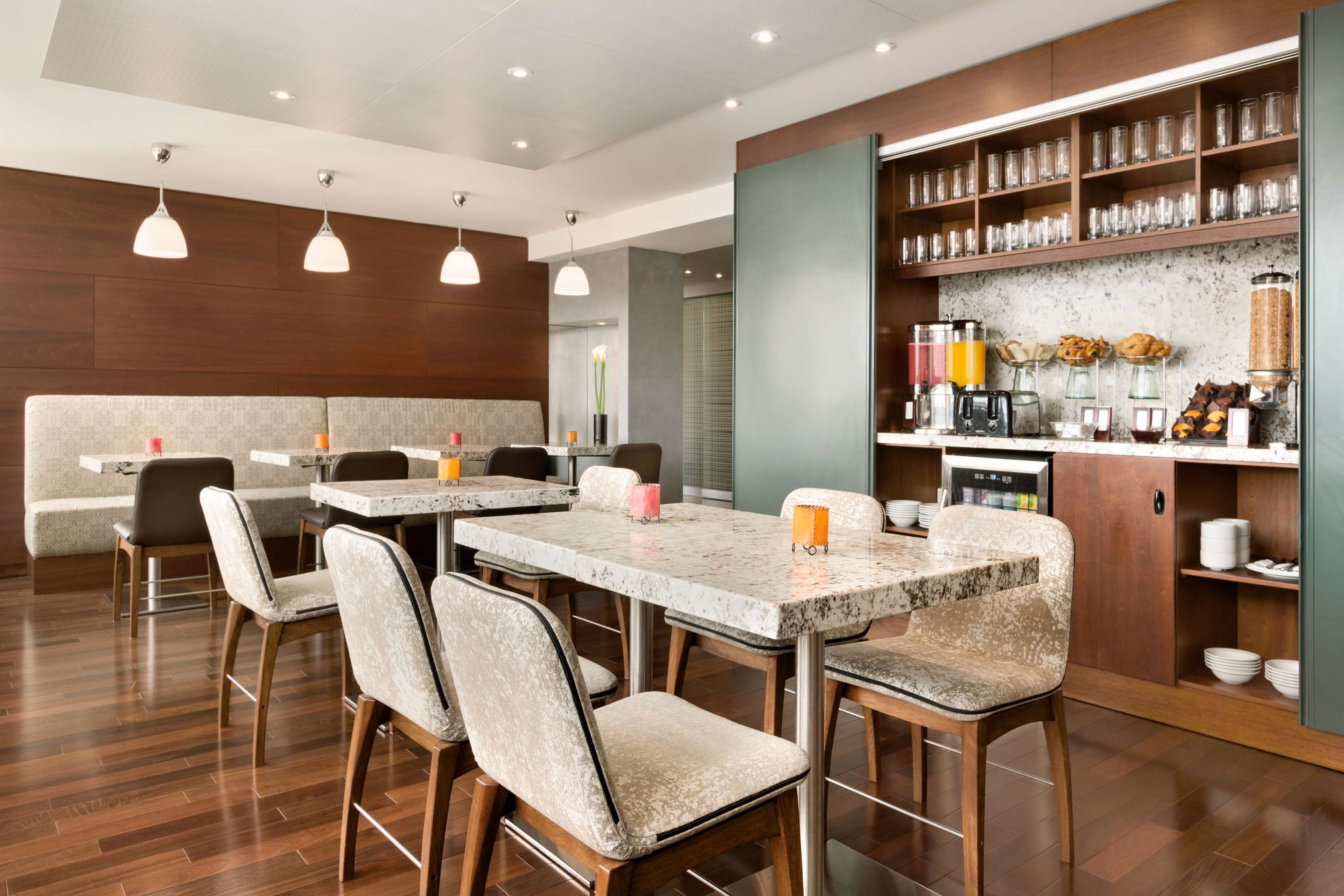 A modern dining area with marble tables and cushioned chairs, a bar with various beverages, and a kitchen area with shelves and appliances.