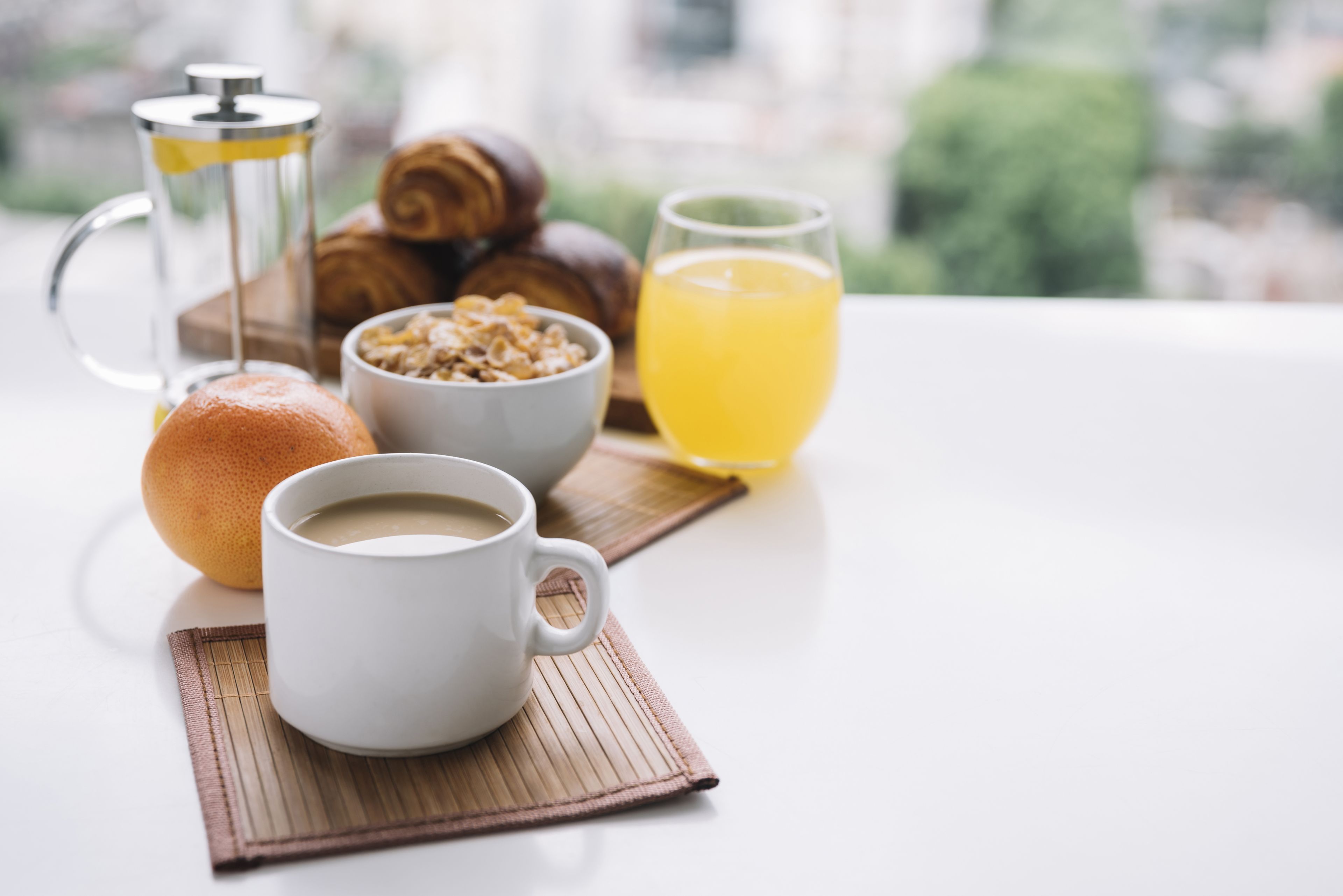 Un desayuno con una taza de café, un vaso de jugo de naranja, un croissant, un muffin y una prensa francesa sobre una mesa.