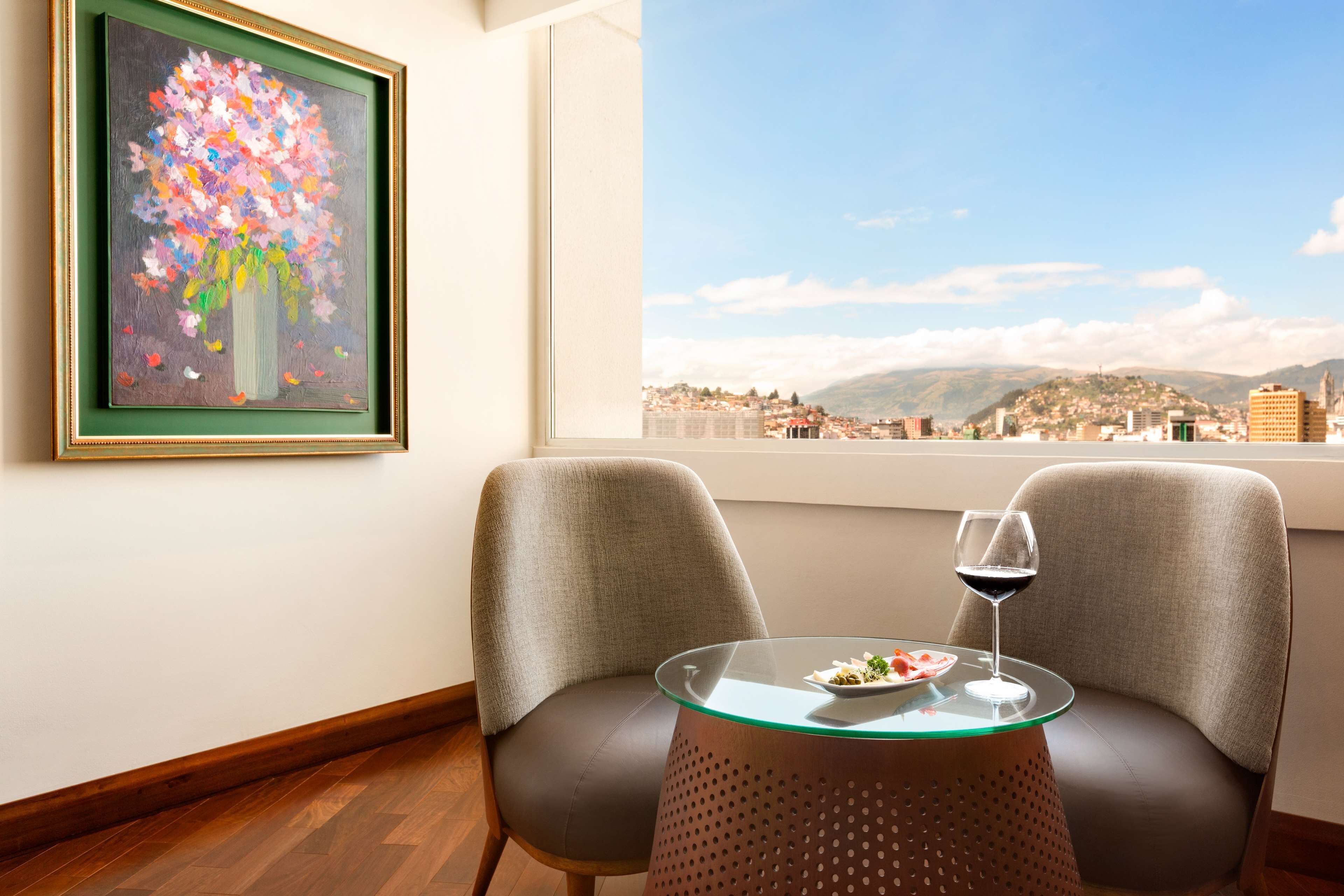 A room with two chairs, a glass table with a plate of food and a wine glass, a framed painting, and a view of a cityscape through a window.