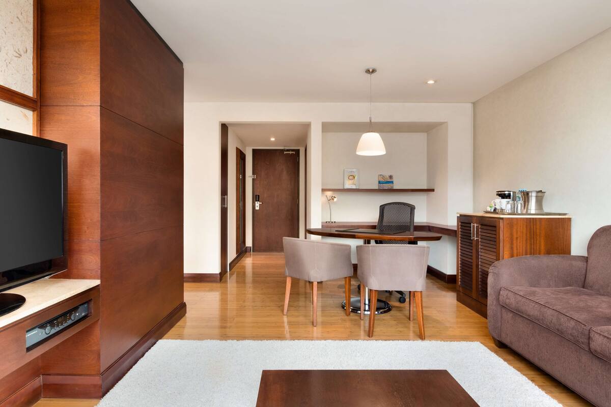 A modern living room with a flat-screen TV, a dining table with chairs, a sofa, and a kitchen area with cabinets and a hanging light fixture.