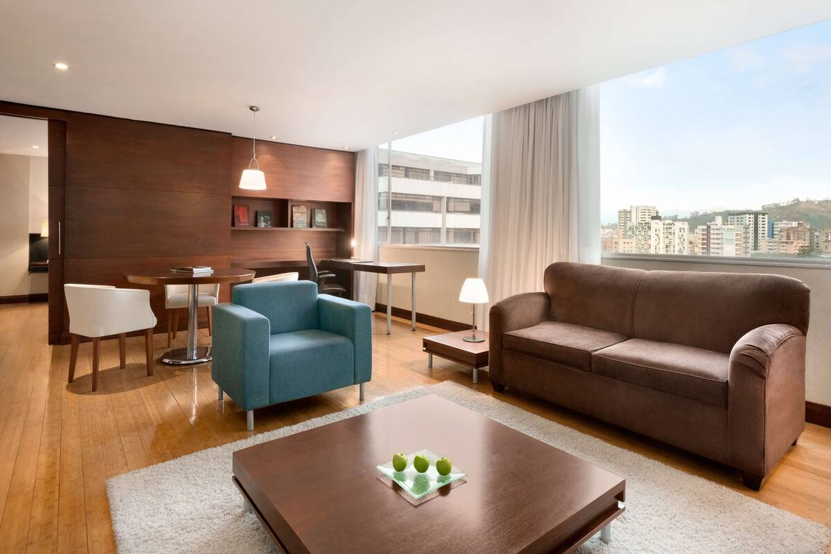 A modern hotel room with a dining area, a sofa, a coffee table, and a large window offering a city view.