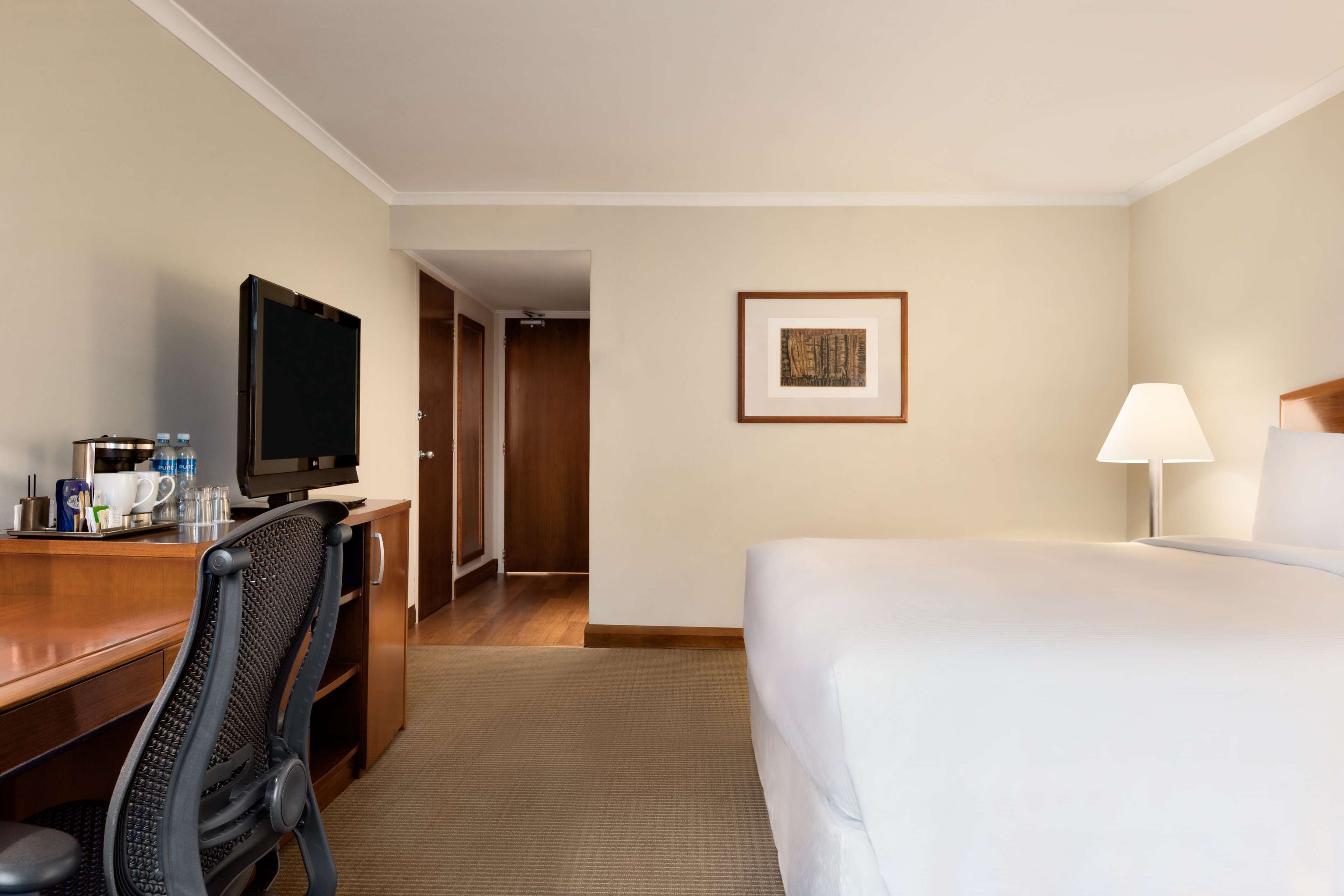 A hotel room with a bed, a desk with a TV, a chair, a lamp, and a framed picture on the wall.