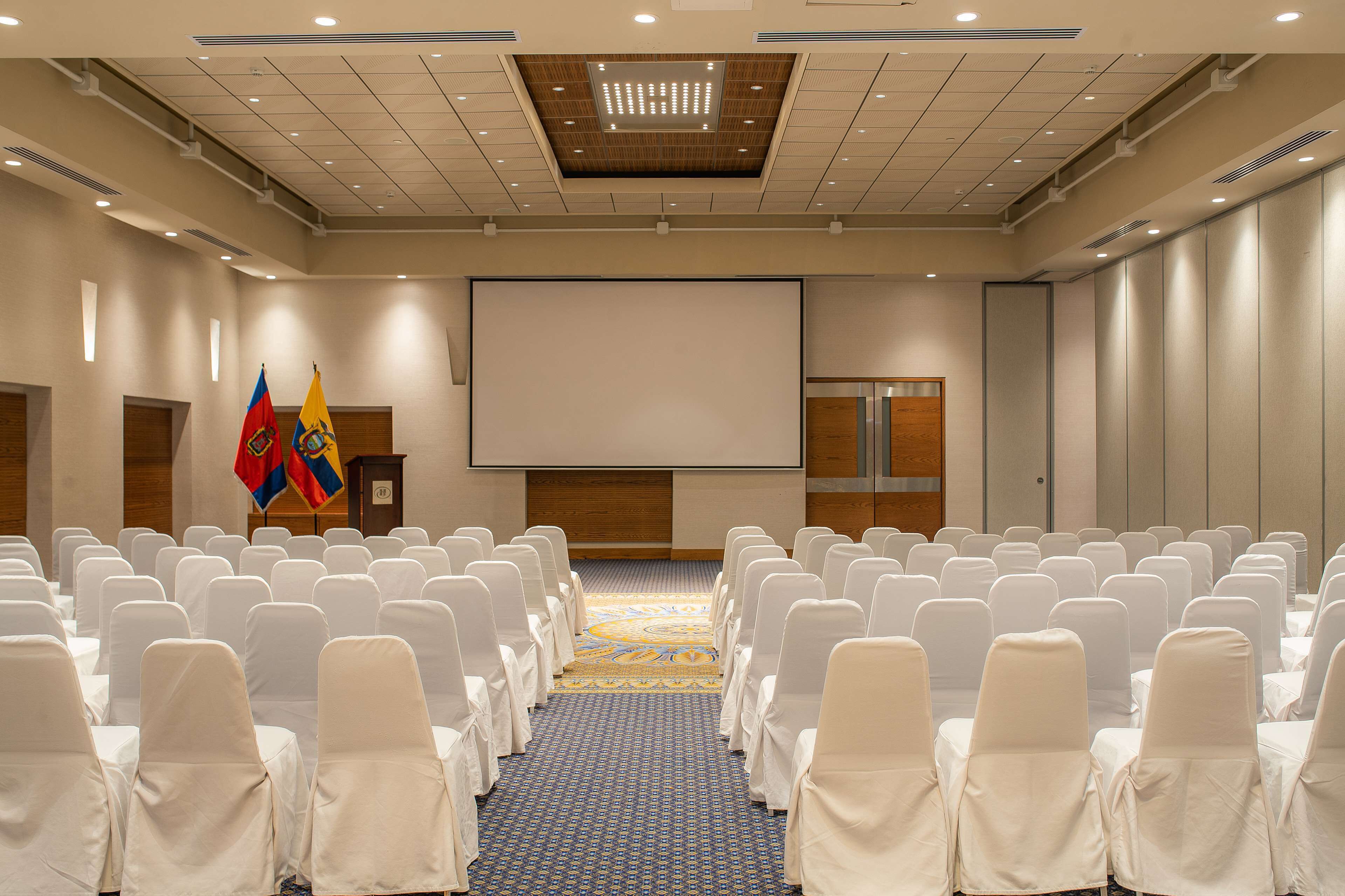 A conference room with rows of white chairs, a large screen, and two flags.