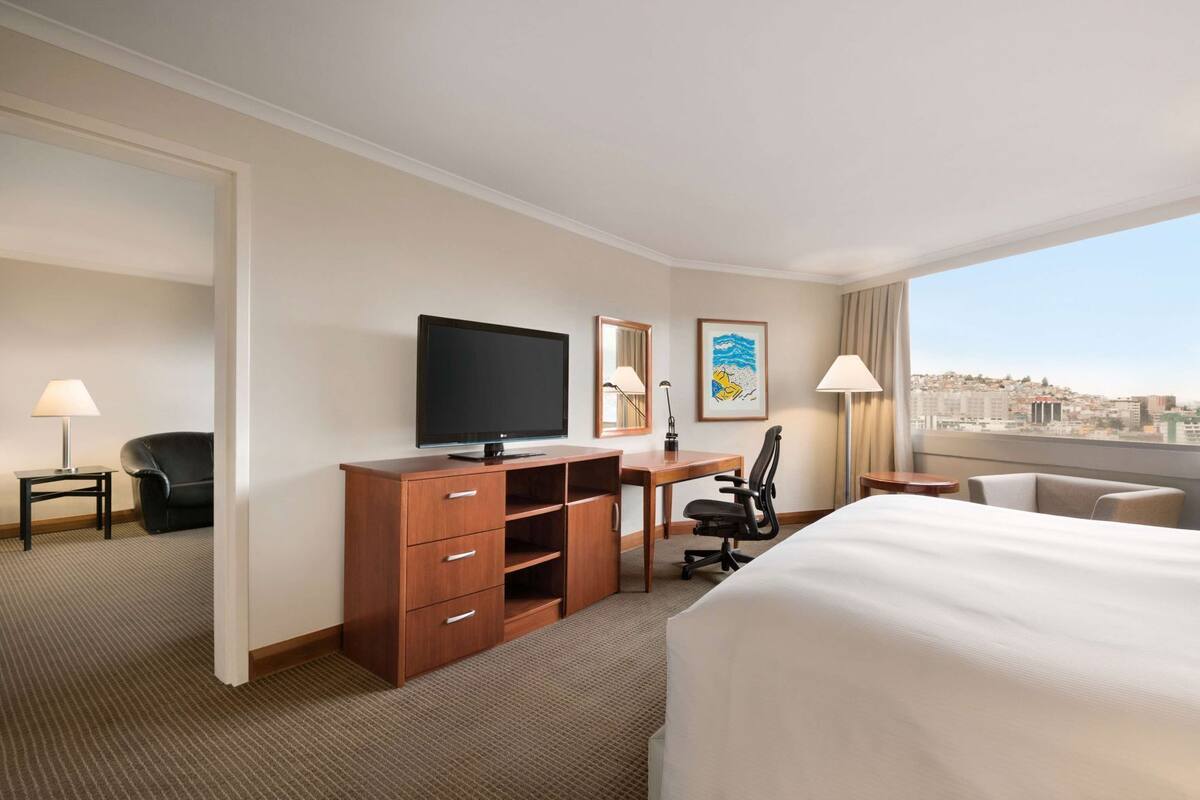 A hotel room with a bed, desk, TV, chair, and city view.