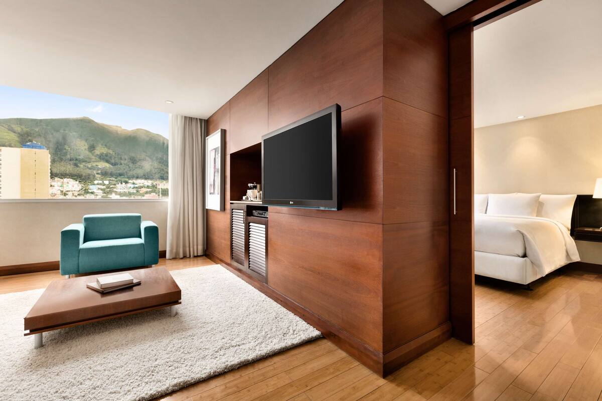 A modern hotel room with a large flat-screen TV mounted on a wooden wall, a teal sofa, a white bed, and a wooden floor.