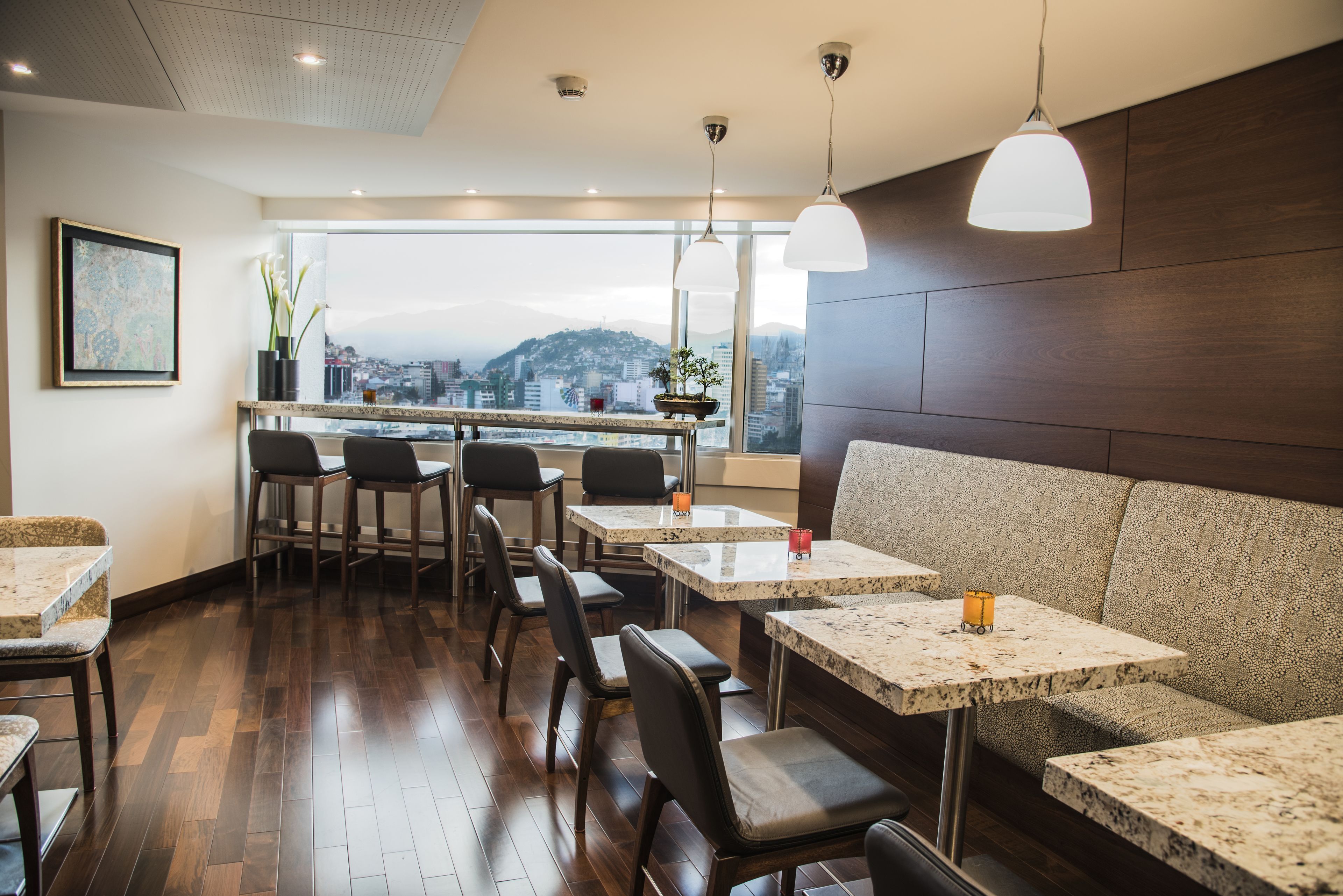 Um restaurante moderno com vista para o horizonte urbano, apresentando um espaço de bar, assentos com mesas e cadeiras, e iluminação pendente.