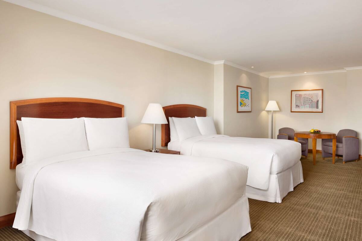 A hotel room with two beds, a desk, two chairs, and a lamp.