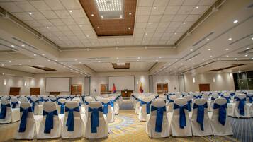 Un salón de conferencias con filas de sillas blancas con cintas azules, una alfombra estampada y una pantalla grande al frente.