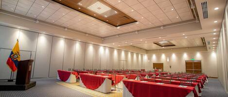 A conference room with rows of tables covered in red tablecloths, a podium with a flag, and a patterned carpet.
