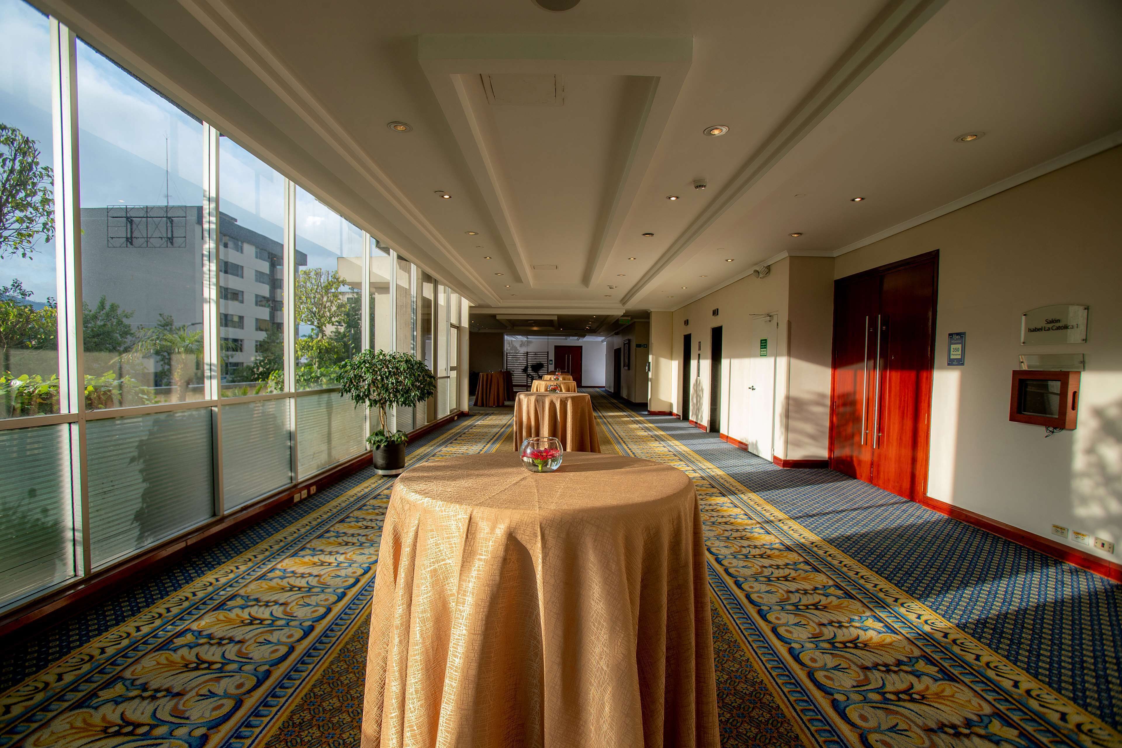 A hotel corridor with a patterned carpet, a table with a vase of flowers, and a view of the outside through large windows.
