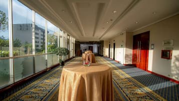 A hotel corridor with a patterned carpet, a table with a vase of flowers, and a view of the outside through large windows.