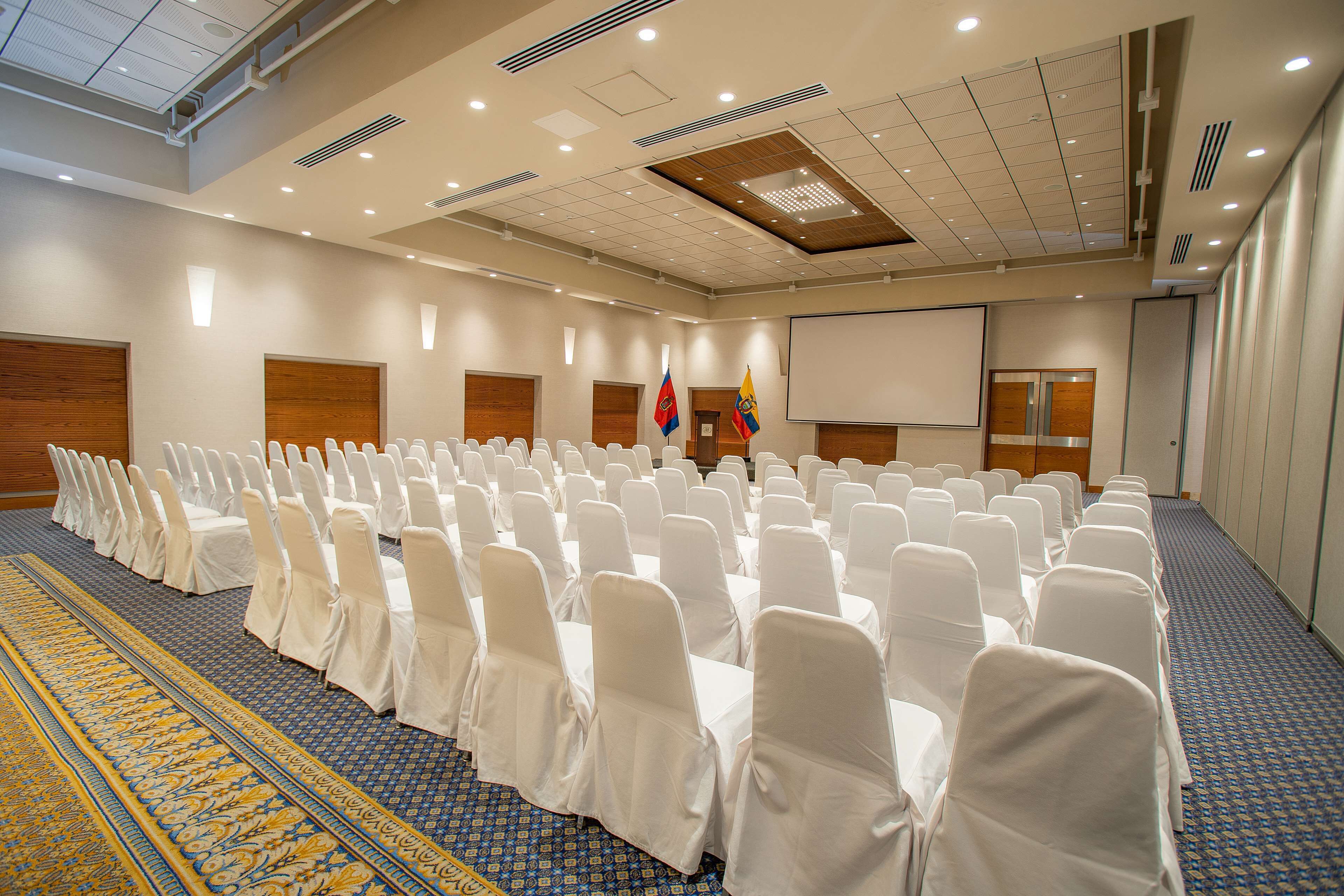Ein Konferenzraum mit Reihen weißer Stühle, einer Projektionsleinwand und Flaggen.