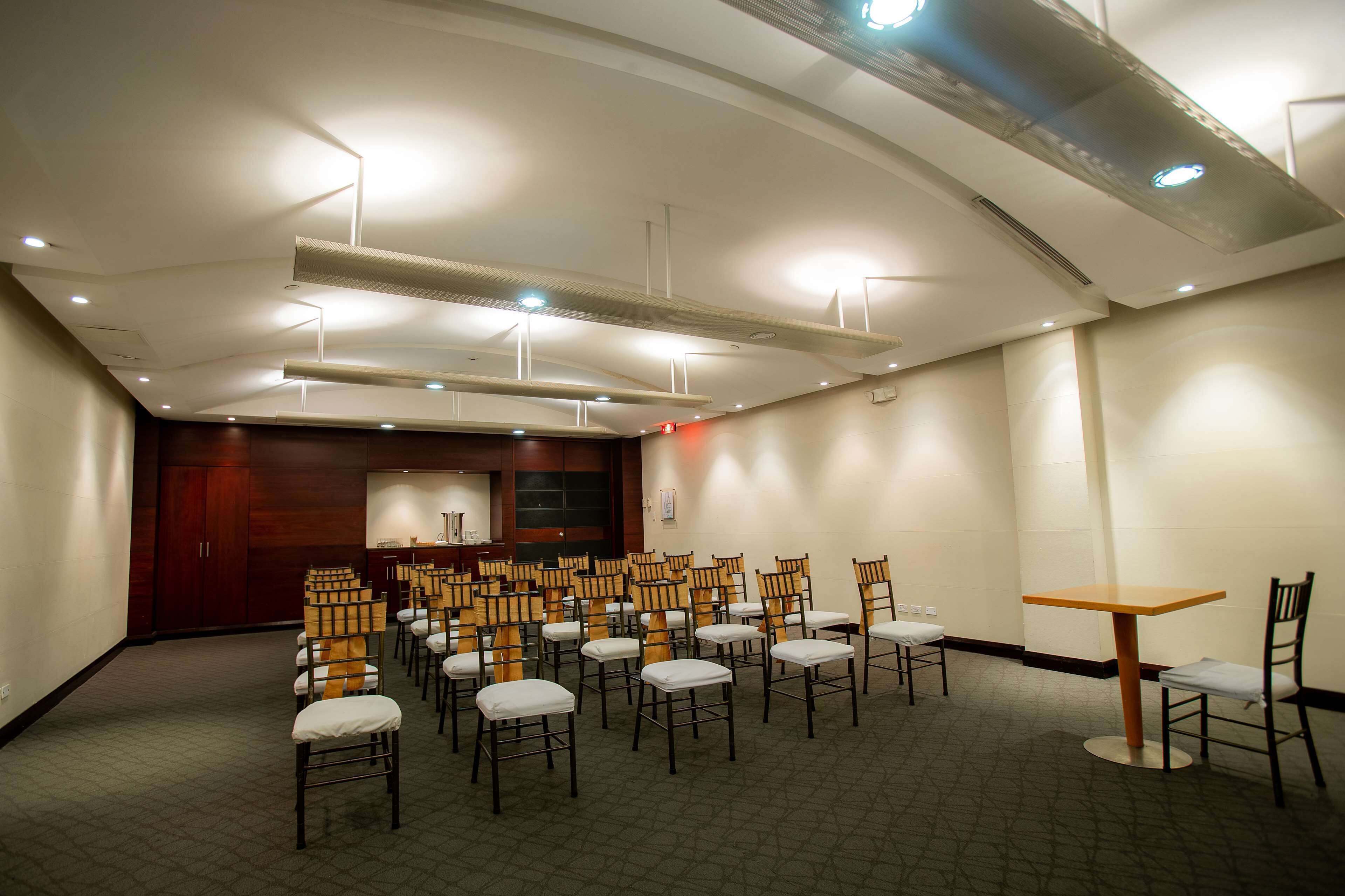 A conference room with rows of chairs and a table, equipped with ceiling lights and a wooden panel wall.