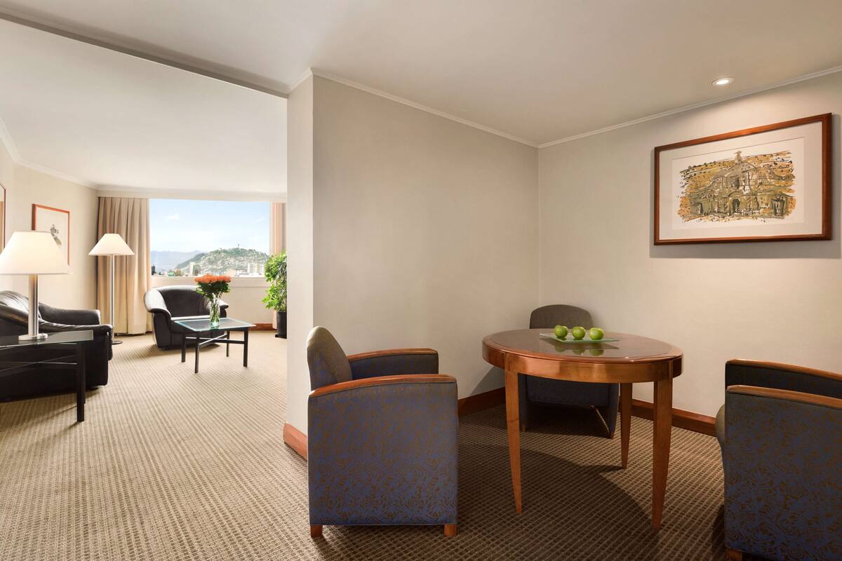 A hotel room with a seating area, a dining table with apples, and a framed picture on the wall.