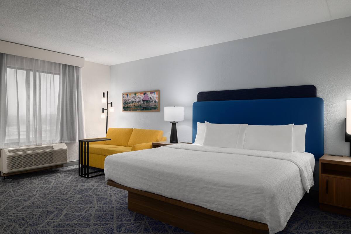 Une chambre d’hôtel avec un grand lit, un fauteuil jaune, un bureau avec une lampe et un tableau au mur.