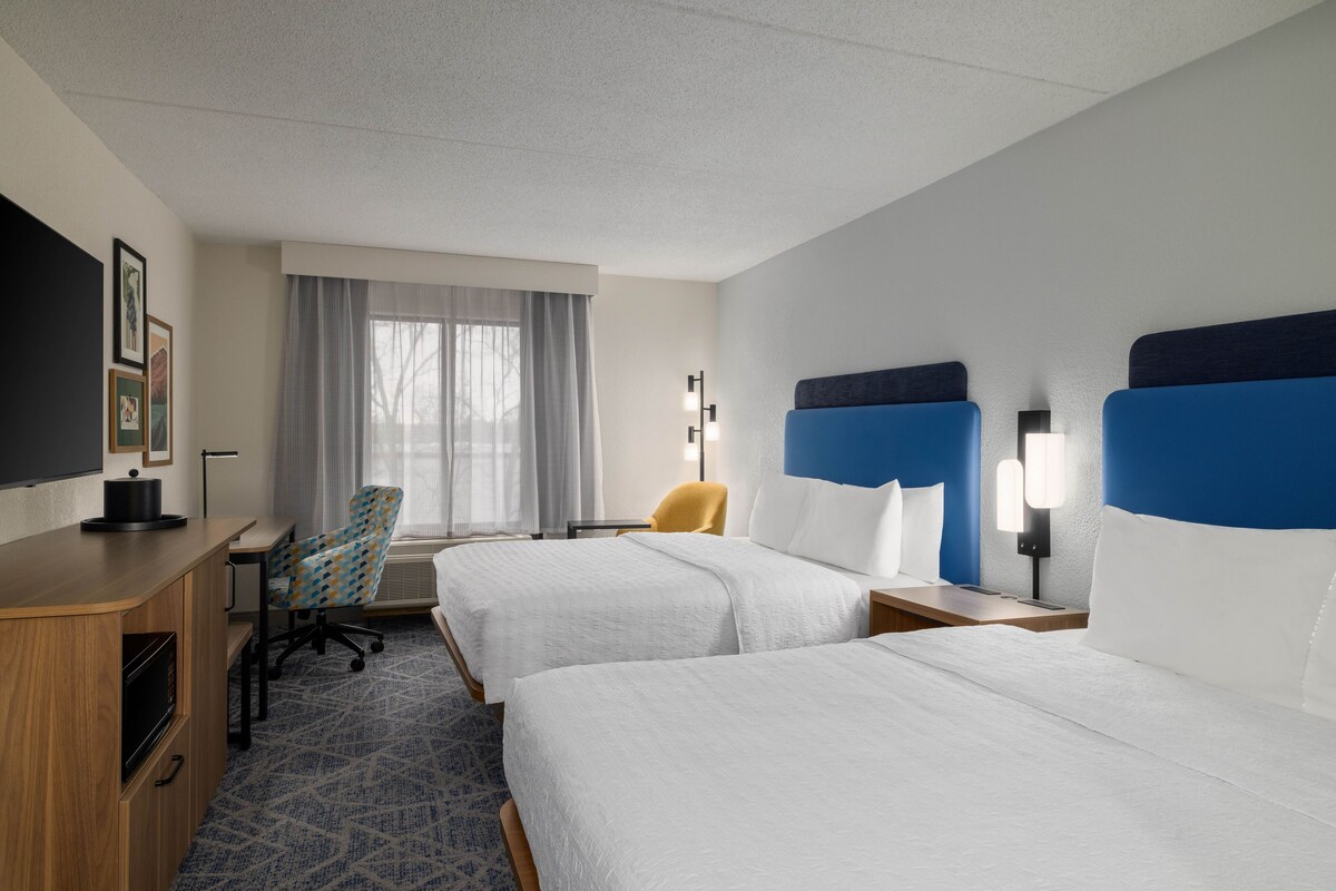 A hotel room with two beds, a desk, a chair, a TV, and a window with curtains.