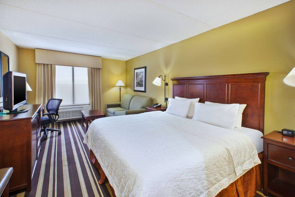 Une chambre d’hôtel avec un grand lit, un bureau avec un ordinateur, une chaise, une lampe et une horloge sur la table de chevet.