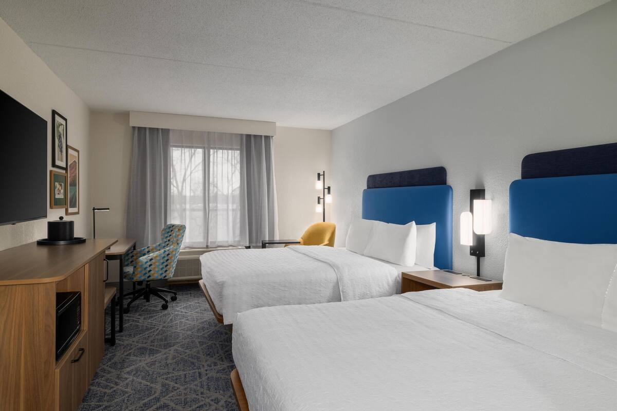 Une chambre d’hôtel avec deux lits, un bureau, une chaise, une télévision et une fenêtre avec des rideaux.
