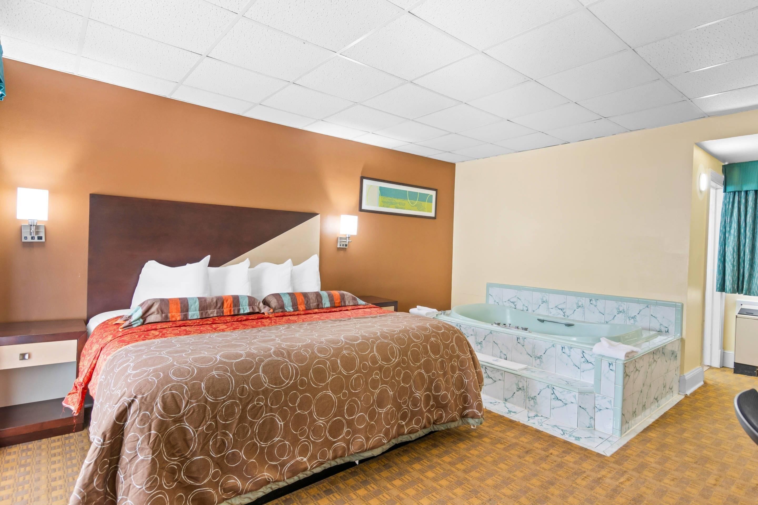 A hotel room with a bed, a nightstand, a wall-mounted lamp, a painting, a bathroom with a bathtub, and a marble-patterned sink.