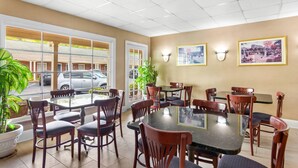 A hotel dining area with round tables and chairs, large windows, and wall-mounted light fixtures. - Rodeway Inn Williamsburg Historic Area (Williamsburg)
