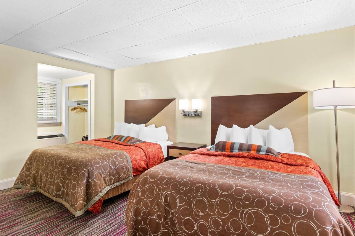 A hotel room with two beds, each with a brown patterned bedspread and white pillows. There is a nightstand with a lamp between the beds, and a window with blinds on the left side.