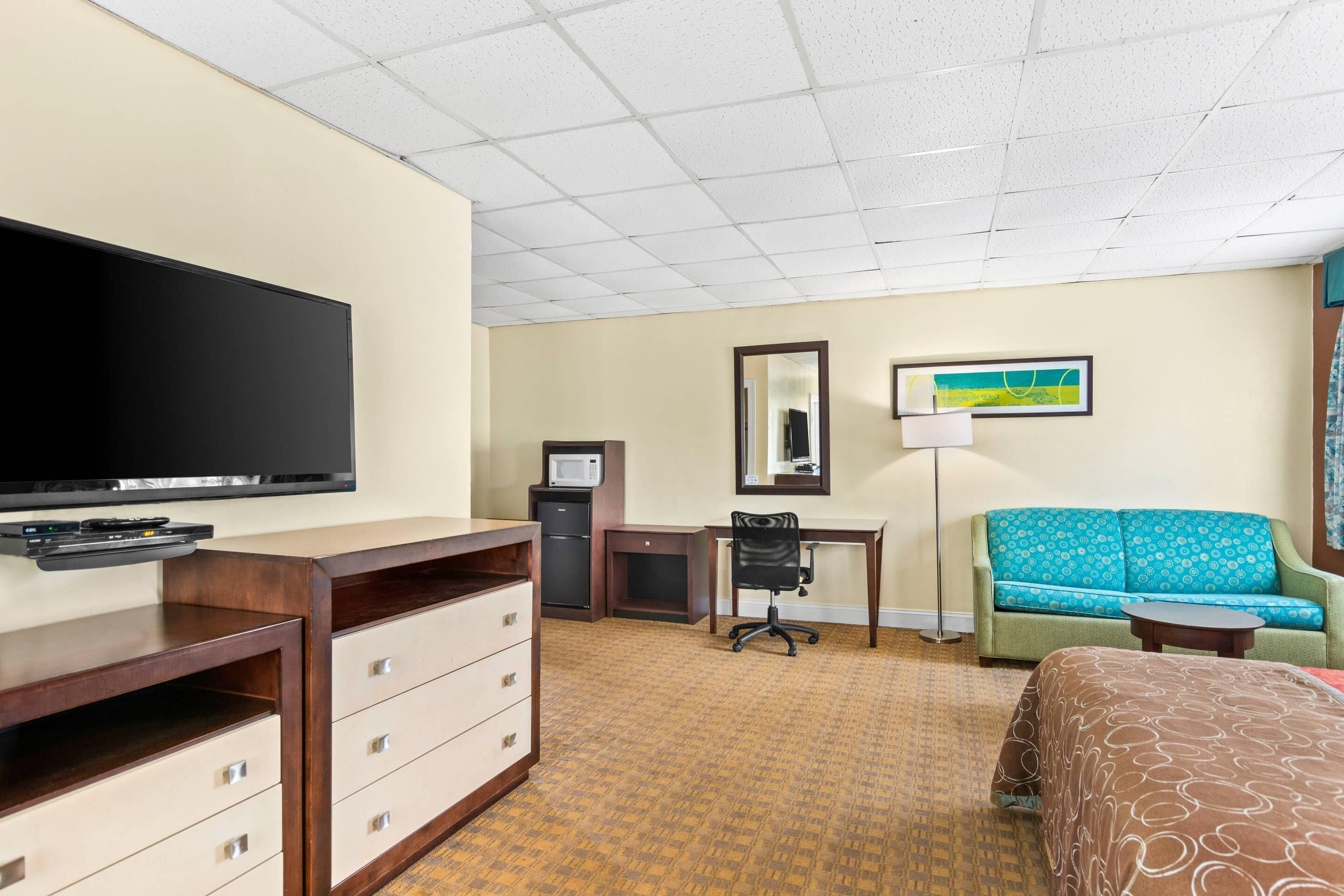 A hotel room with a flat-screen TV, a desk with a computer, a green sofa, a bed, and a small table.