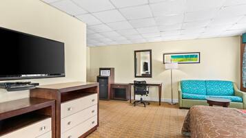 A hotel room with a flat-screen TV, a desk with a computer, a green sofa, a bed, and a small table.