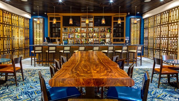 A bar area with a long wooden table, multiple bar stools, and a well-stocked liquor shelf. - DoubleTree by Hilton Hotel Arlington DFW South (Arlington)