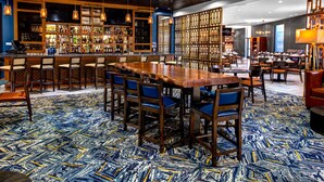 A bar area with a long wooden table and chairs, a bar counter with bottles, and a patterned carpet. - DoubleTree by Hilton Hotel Arlington DFW South (Arlington)
