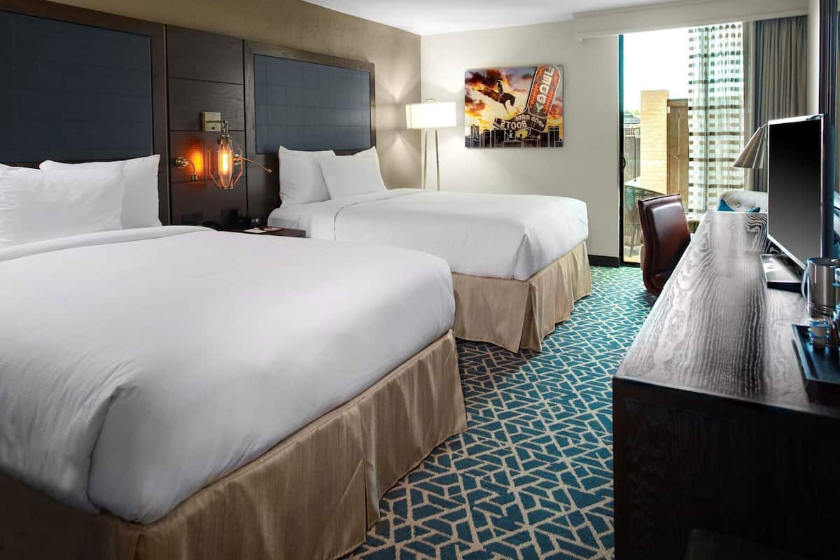 A hotel room with two beds, a desk, a chair, a TV, and a cityscape painting.