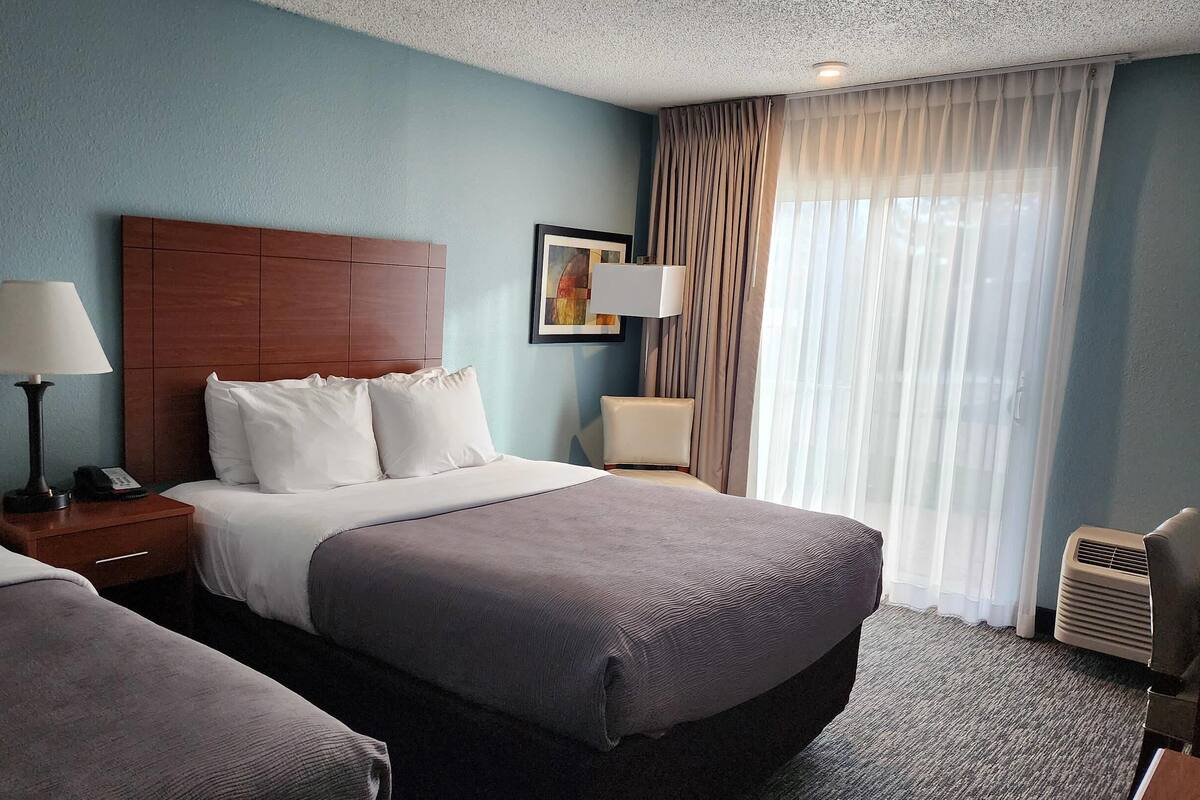 A hotel room with two beds, a wooden headboard, a nightstand with a lamp, a bedside table with a lamp, a framed picture on the wall, and a window with sheer curtains.