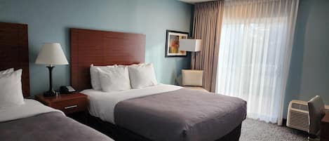 A hotel room with two beds, a wooden headboard, a nightstand with a lamp, a bedside table with a lamp, a framed picture on the wall, and a window with sheer curtains.