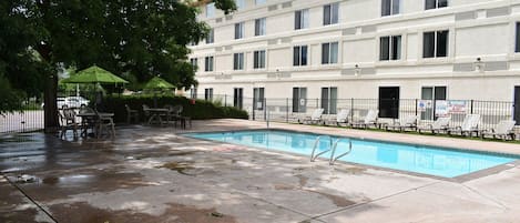 Zona de piscina con sillas de descanso y sombrillas, rodeada por un edificio con múltiples ventanas y unidades de aire acondicionado.