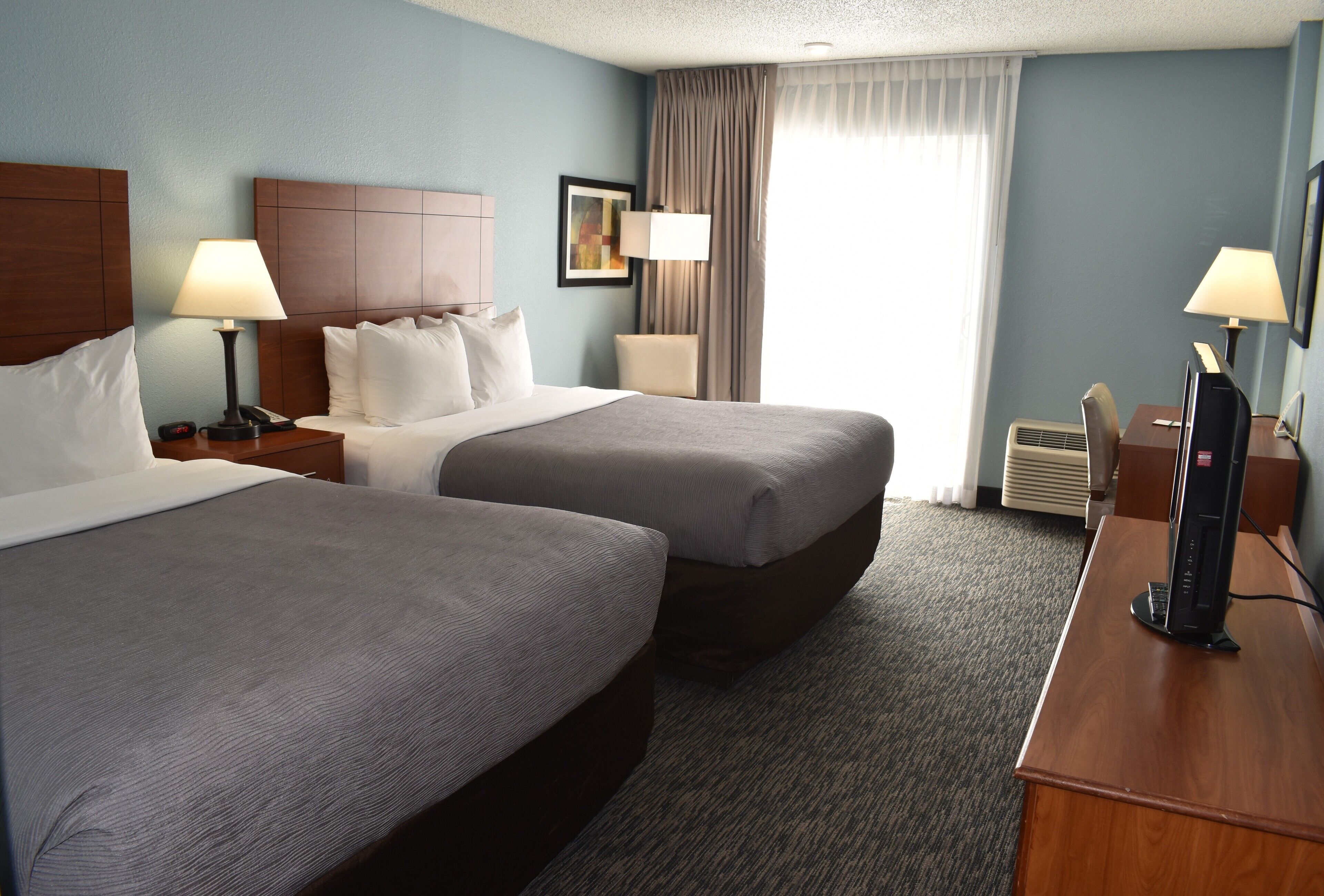 Quarto de hotel com duas camas, uma mesa, uma cadeira, uma televisão e uma janela com cortinas.