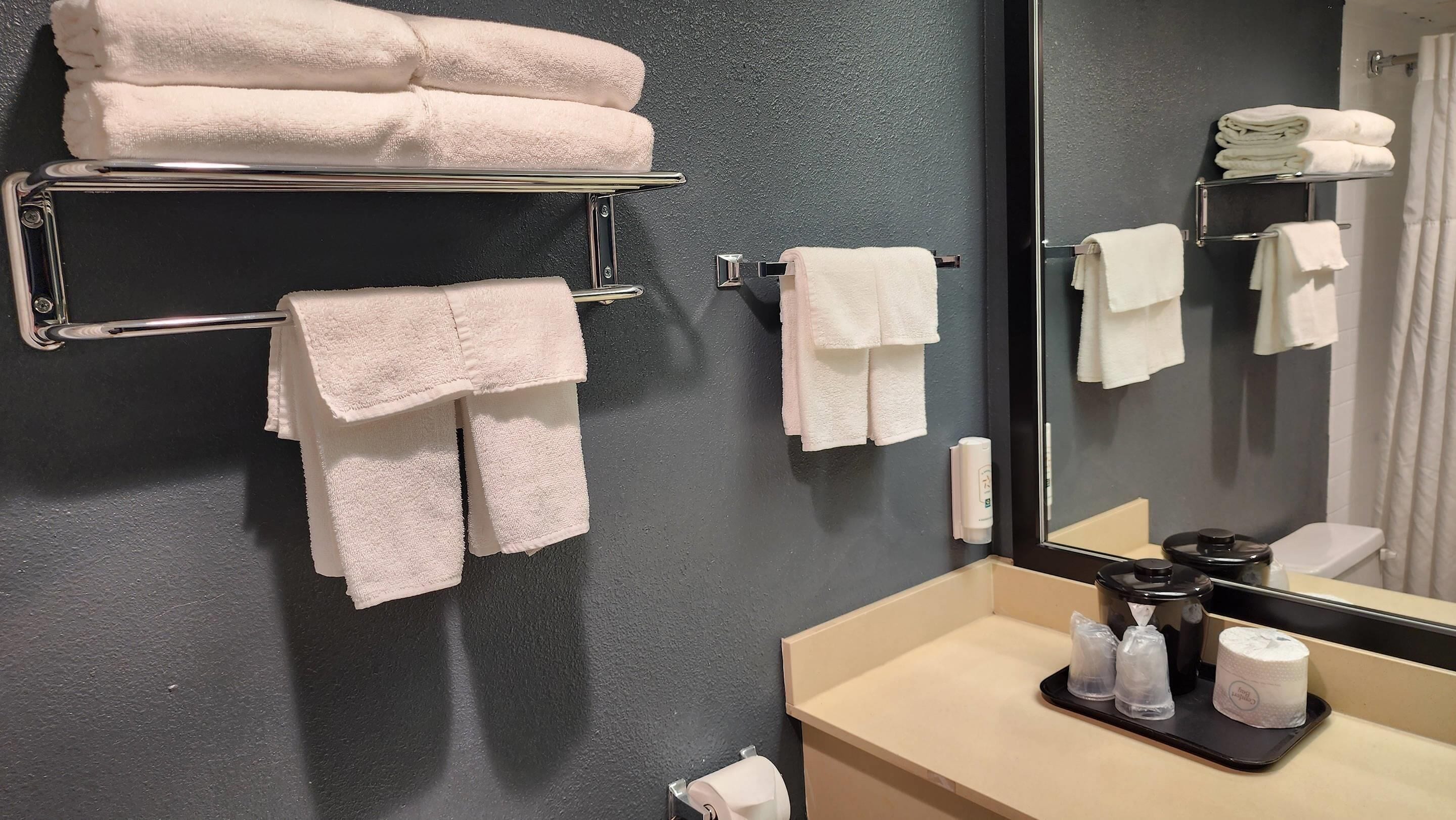 Un baño con pared oscura, un espejo, un lavamanos y varios toalleros con toallas dobladas.