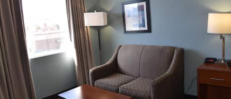 Une chambre d’hôtel avec un canapé, une petite table en bois, une lampe, un tableau encadré au mur et une fenêtre avec des rideaux.