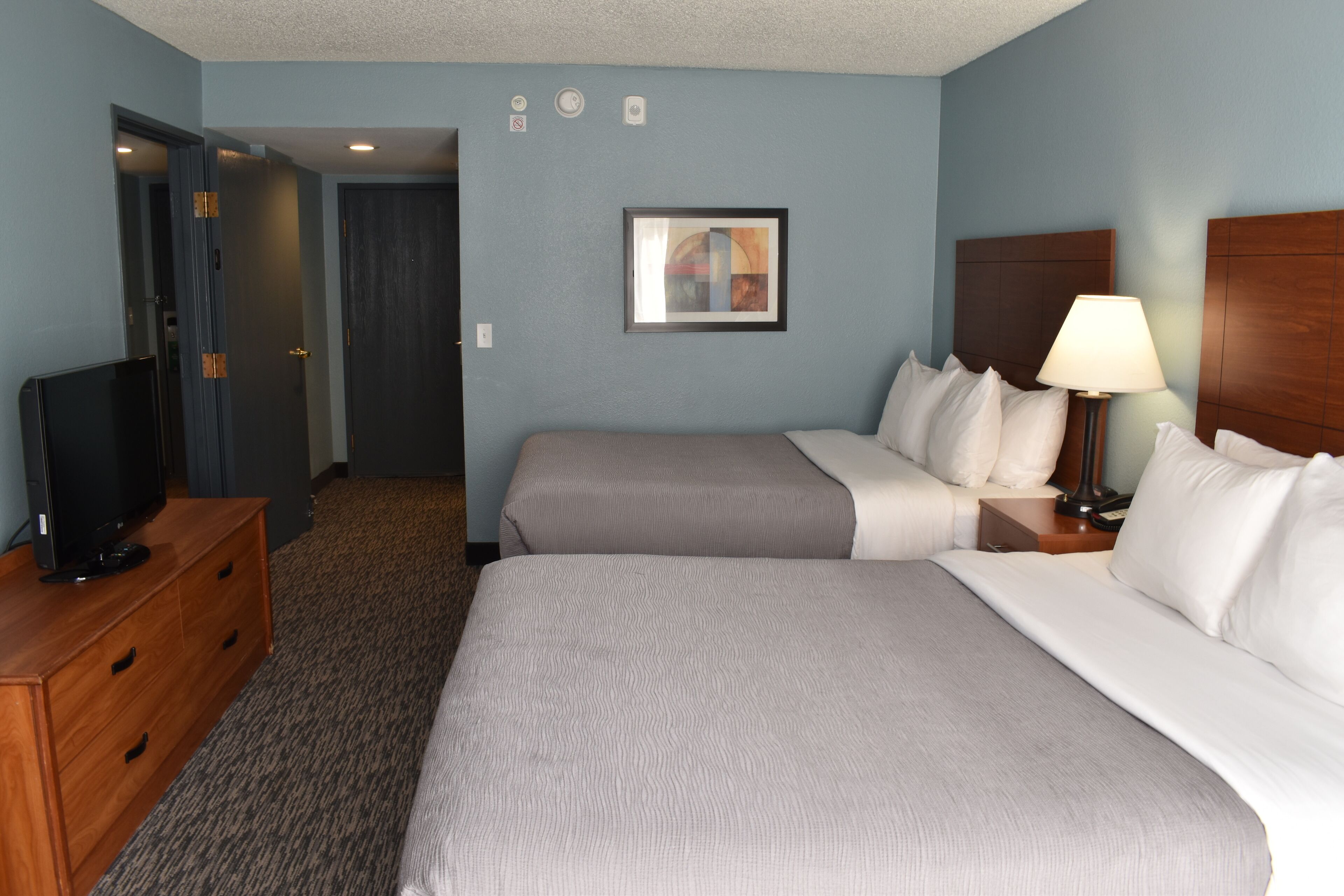 A hotel room with two beds, a television on a dresser, a painting on the wall, and a lamp on the nightstand.