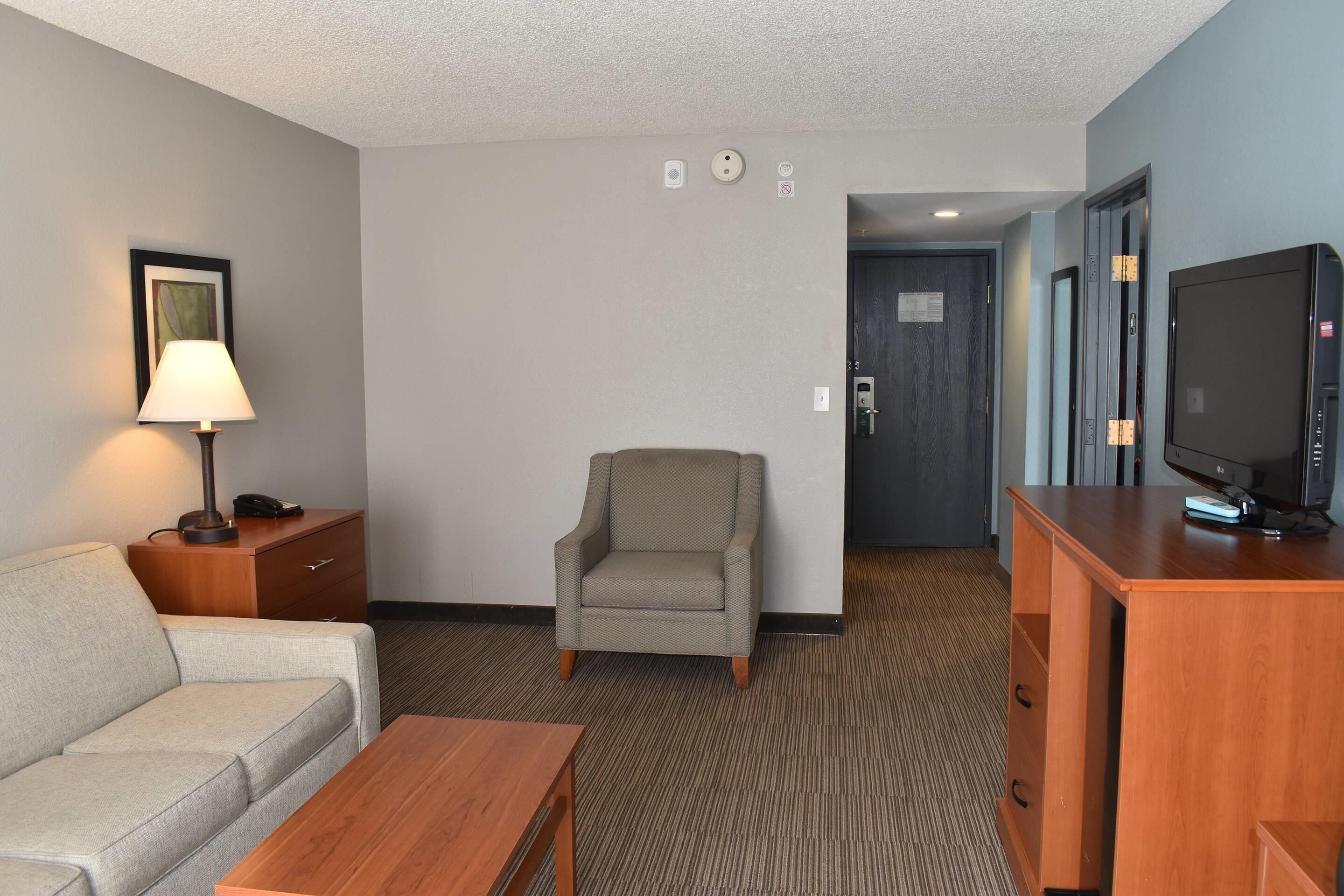 A hotel room with a sofa, armchair, wooden desk, television, and a lamp.