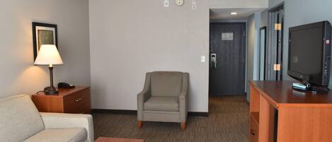 A hotel room with a sofa, armchair, wooden desk, television, and a lamp.