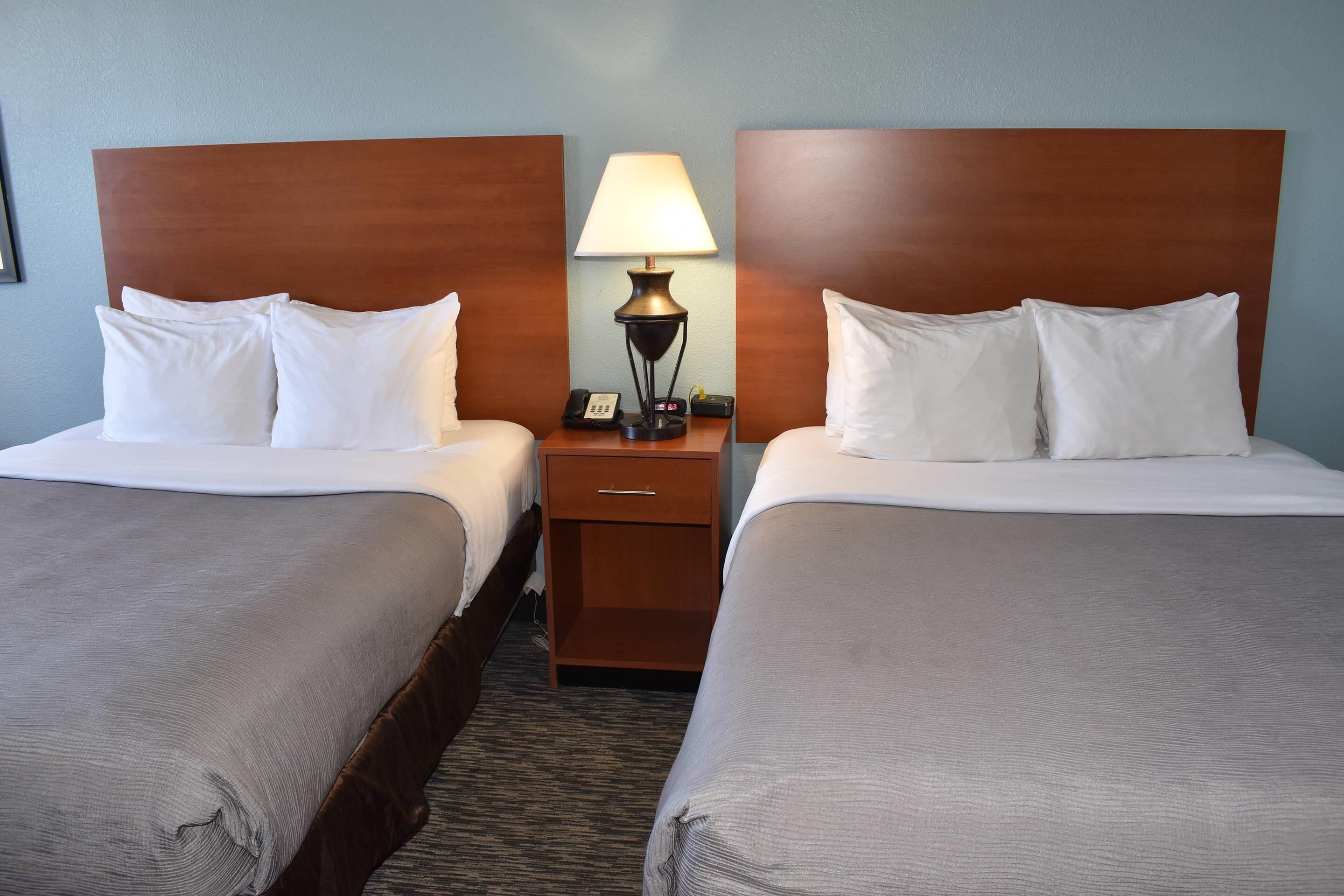 Habitación de hotel con dos camas, cada una con ropa blanca y cabeceros de madera. Un mesita de noche entre las camas contiene una lámpara y un teléfono.