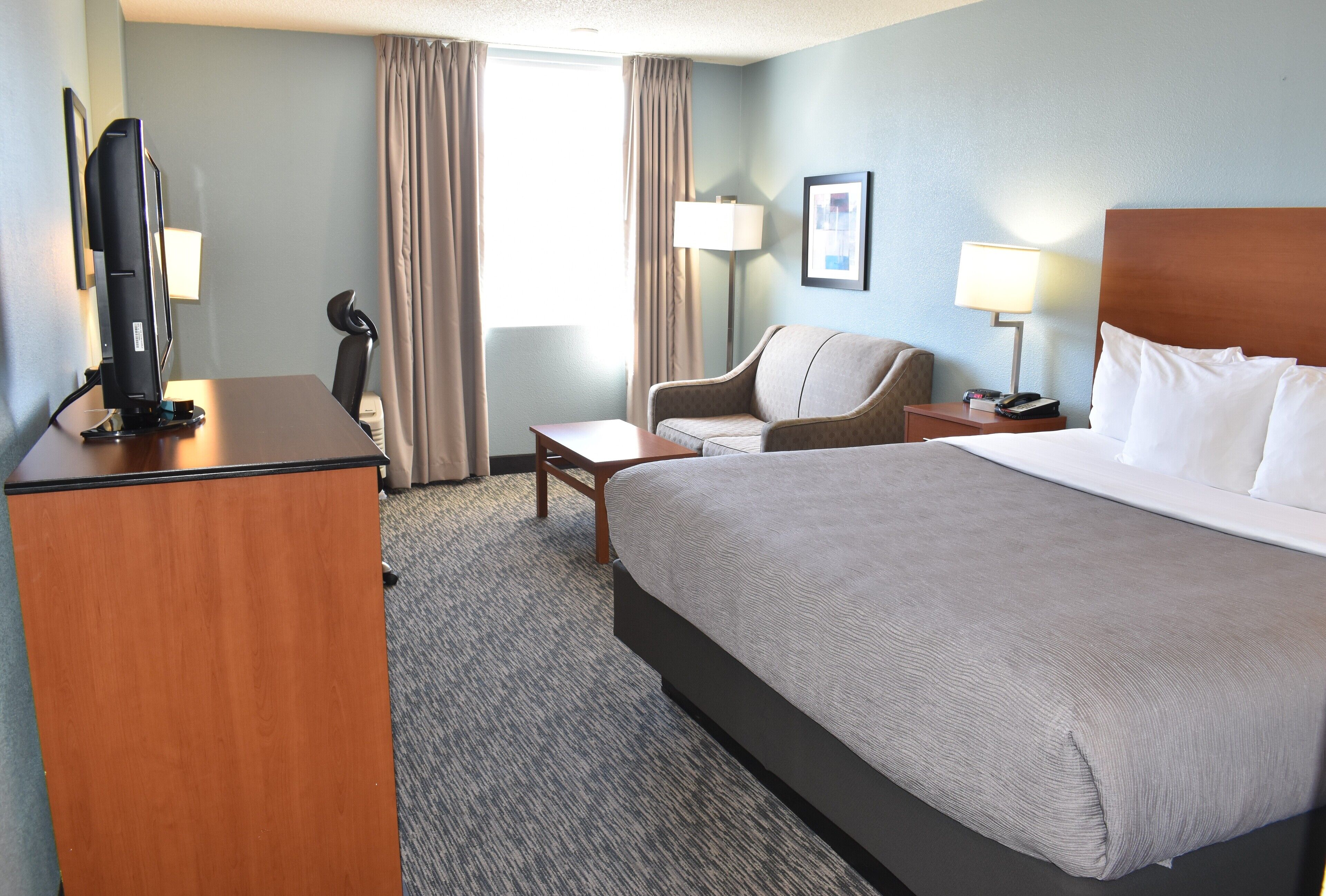 Een hotelkamer met een groot bed, een bureau met een computer, een stoel, een nachtkastje met een lamp en een raam met gordijnen.